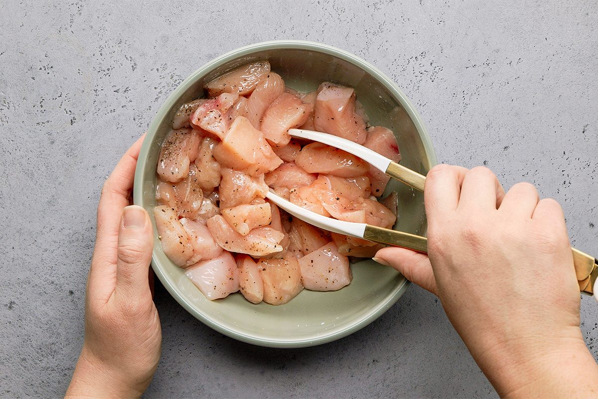 Hands mixing seasoned raw chicken pieces in a green bowl using tongs on a gray speckled surface.