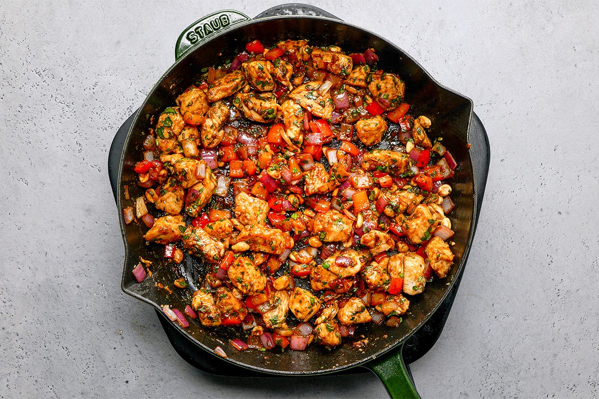 A cast iron skillet filled with cooked diced chicken, red bell peppers, onions, and herbs, all sautéed and seasoned, resting on a gray countertop.
