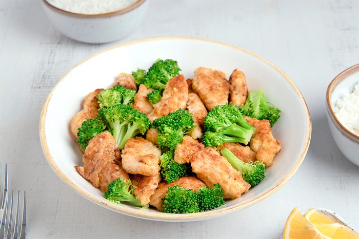 A white bowl filled with pieces of cooked chicken and bright green broccoli florets. The background shows a lemon wedge and small bowls, one possibly containing rice and another with a creamy sauce.