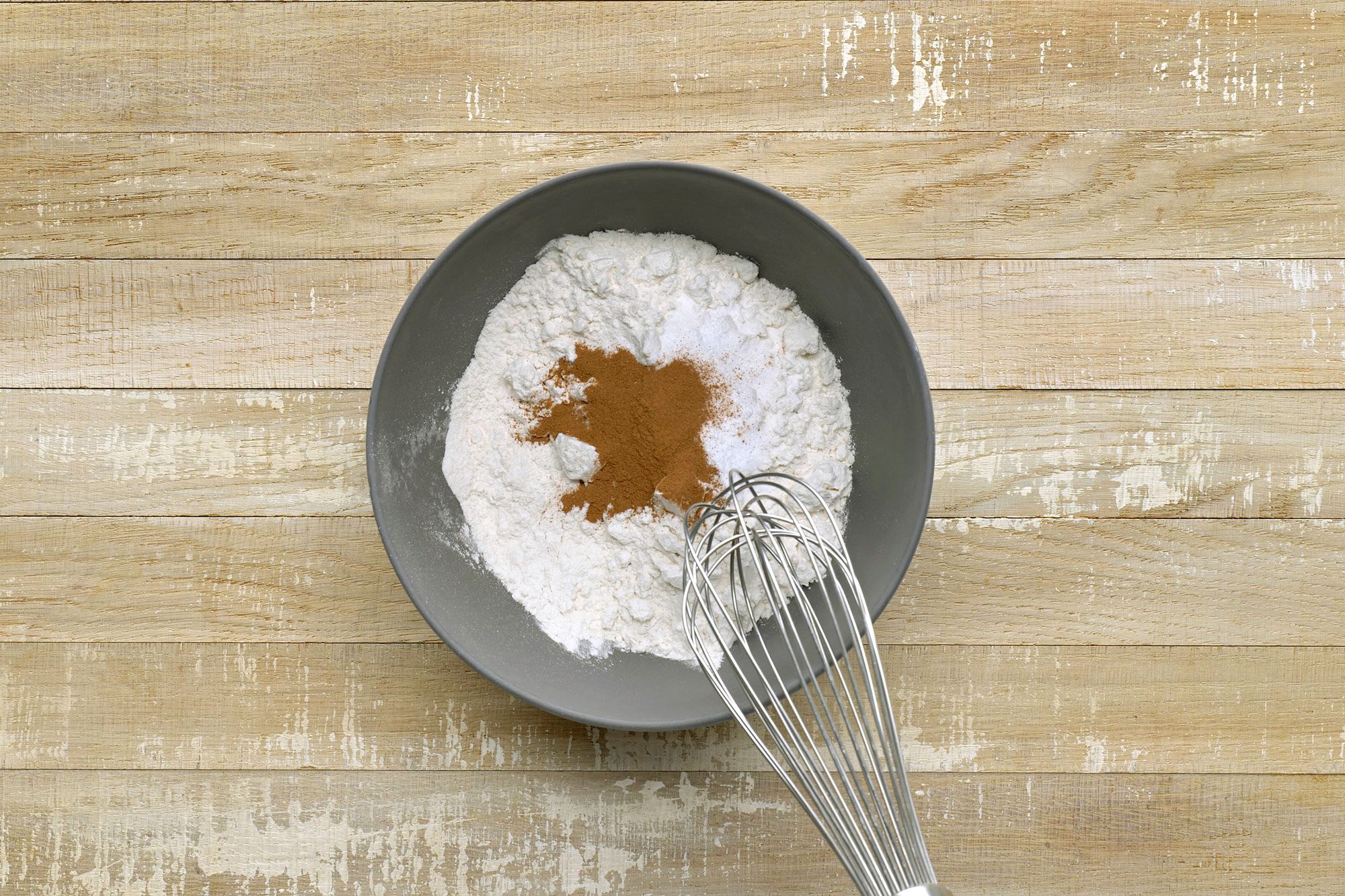 Overhead shot of combine the flour; baking soda; salt and cinnamon; whisk tool; wooden surface;