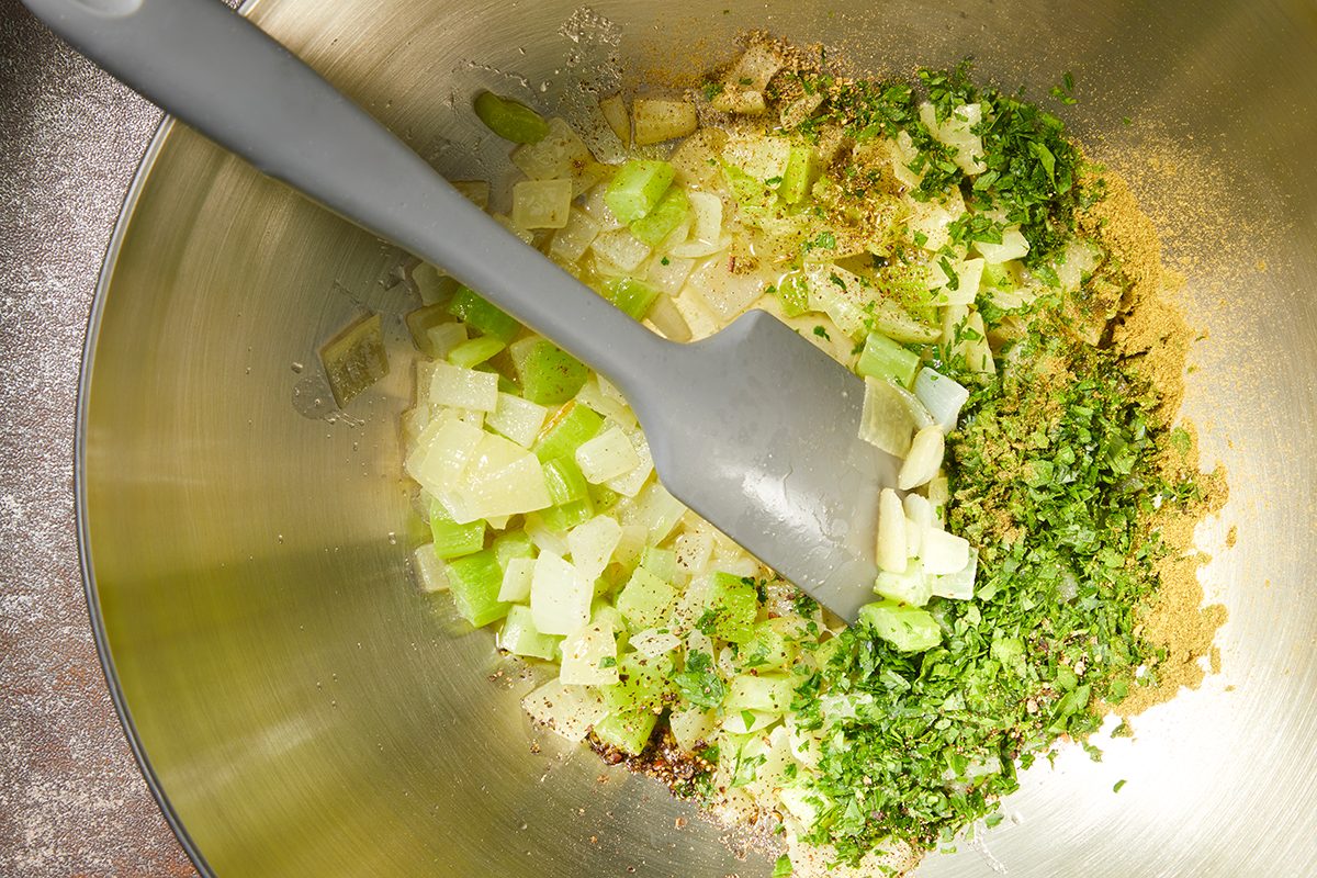 Chopped onions, celery, and parsley in a metal mixing bowl with seasonings, being stirred by a gray spatula.