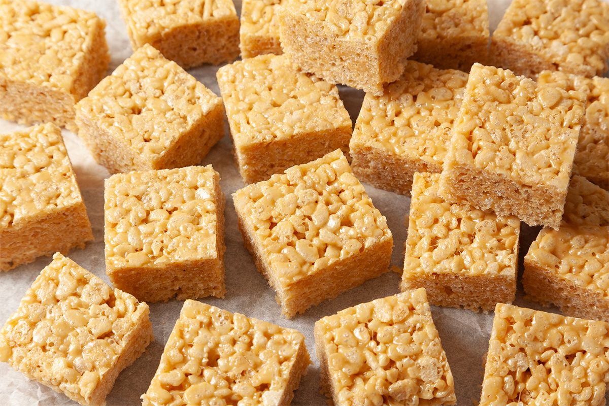 Square pieces of rice crispy treats are arranged on a white surface. They have a golden, crunchy texture from the puffed rice and marshmallow mixture, creating a sweet, chewy appearance.