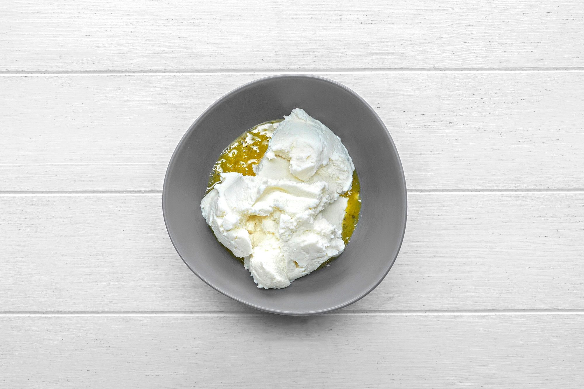 overhead shot of a gray bowl filled with a white, creamy substance, likely ricotta cheese or cream cheese, sitting on a white wooden surface; the bowl is positioned in the center of the image and the wooden surface fills the background