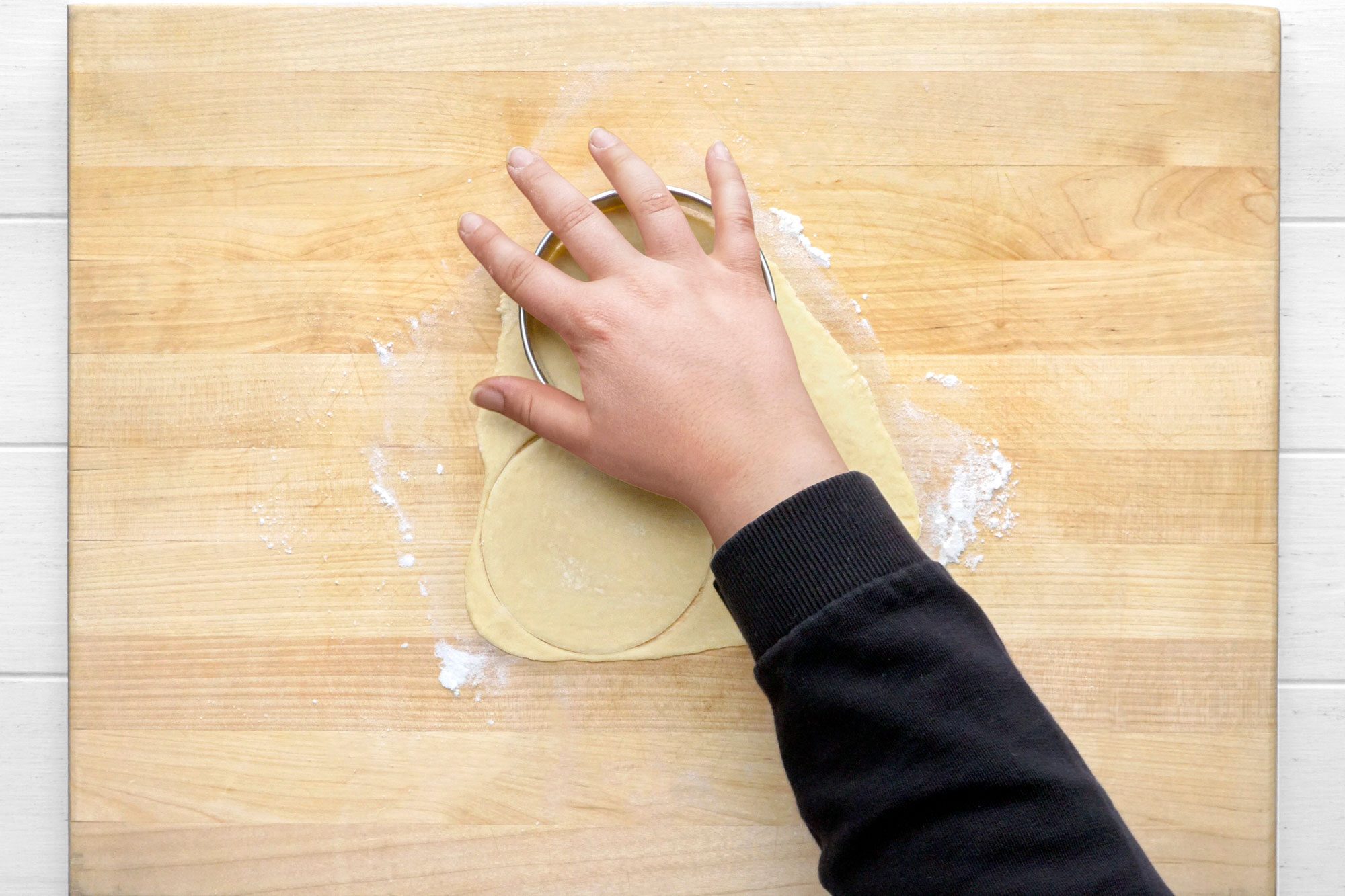 overhead shot of a hand using a round cookie cutter to cut out a circle of dough on a wooden cutting board, the board is covered in a thin layer of white flour
