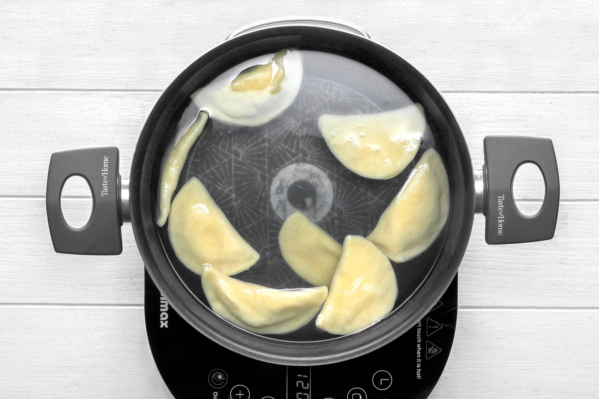 overhead shot of a black pot with a silver rim sits on a black electric stovetop; the pot is filled with water and eight half-moon shaped dumplings are boiling in the water;
