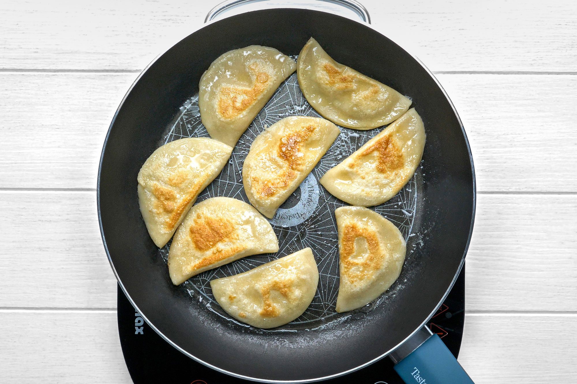 overhead shot of a black frying pan with a blue handle, sitting on a white wooden surface; the frying pan is filled with eight golden-brown dumplings that are cooking in oil, and they are arranged in a circular pattern