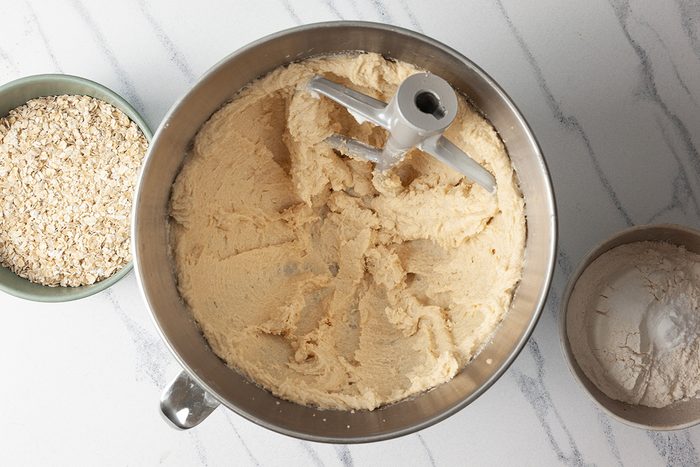 A mixing bowl filled with creamy dough, equipped with a paddle attachment, sits on a marble countertop. To the left, a bowl of oats is visible, and to the right, a bowl of flour.