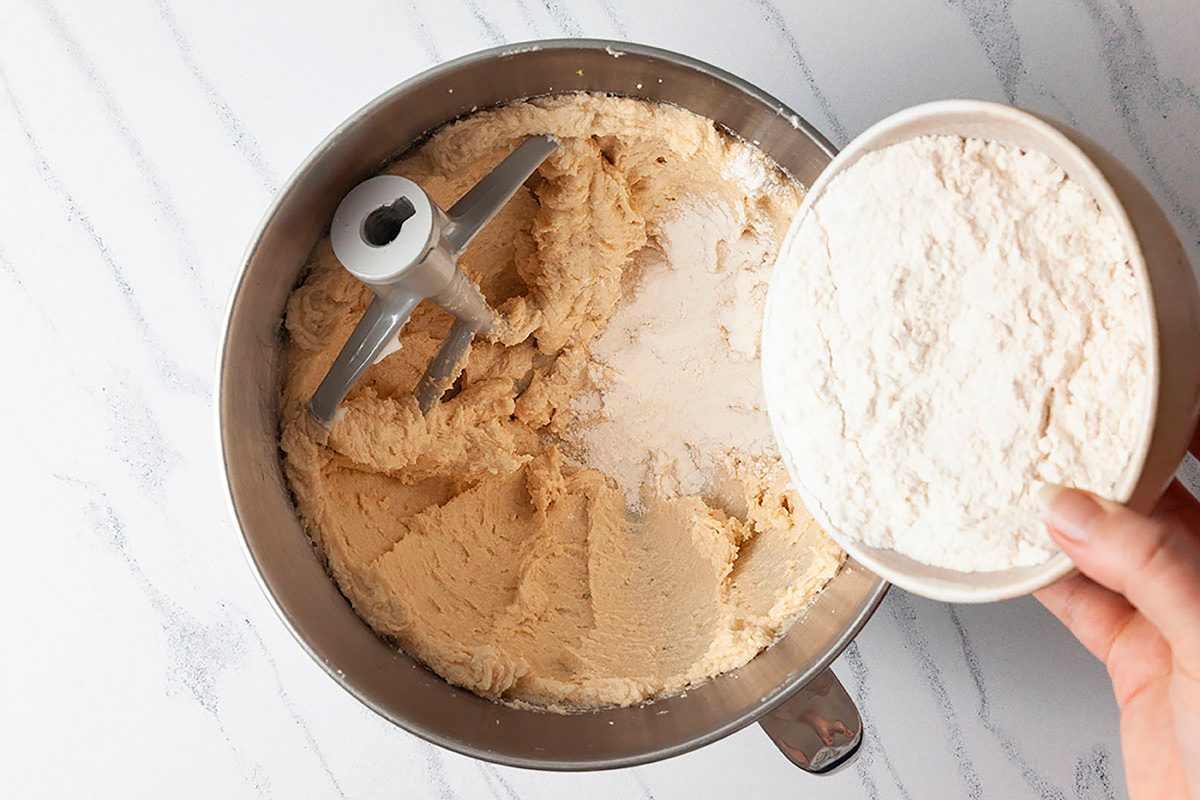 A mixing bowl contains creamy batter, with a paddle attachment visible. A hand is holding a white bowl filled with flour over the batter, seemingly ready to add it in. The background is a light-colored marble surface.