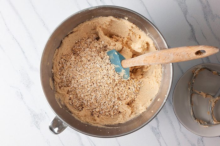 A metal mixing bowl on a marble countertop contains cookie dough with oats being mixed using a blue spatula.