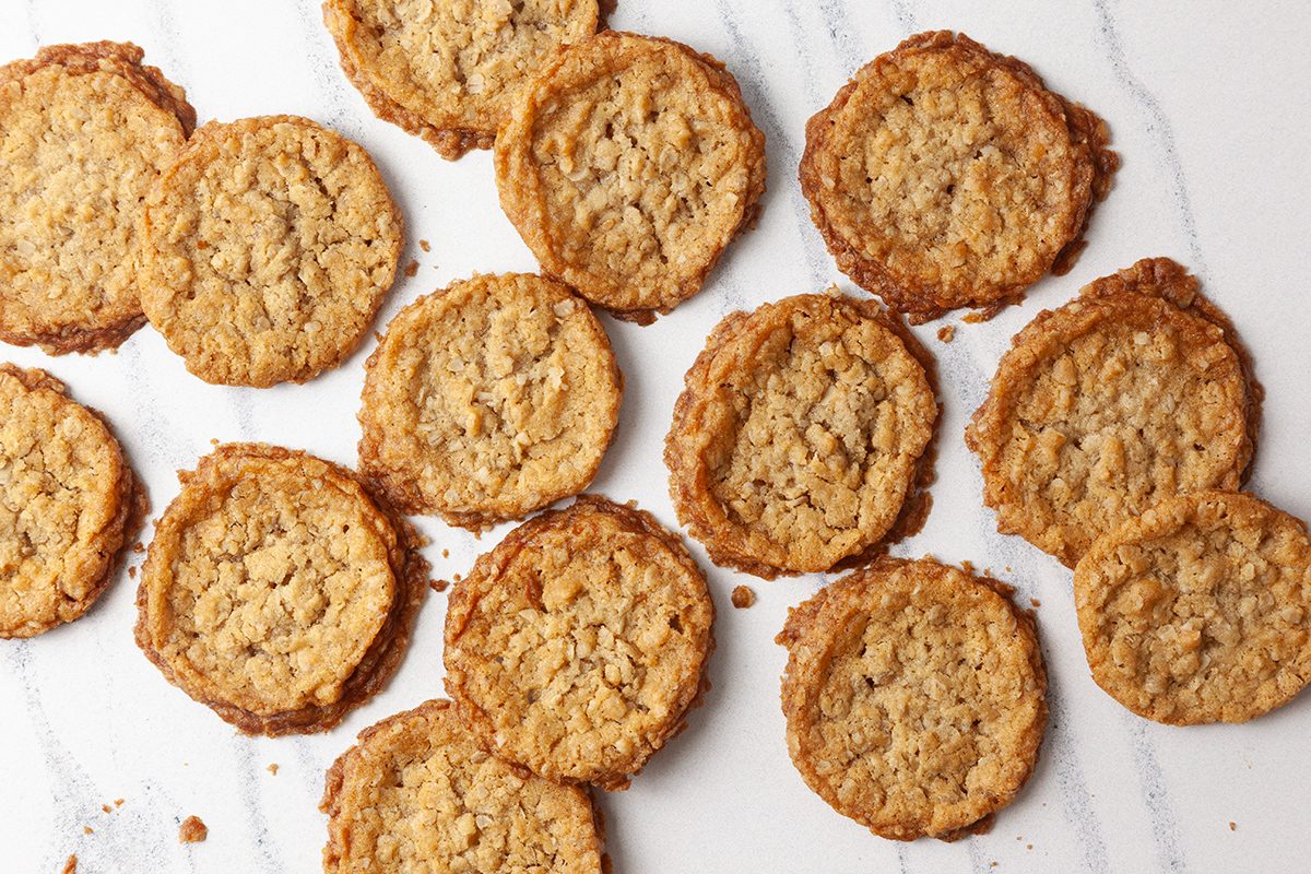 A scattering of round, golden-brown cookies on a white marble surface. The cookies have a textured appearance, likely oatmeal or similar ingredients. The arrangement is informal and slightly overlapping.
