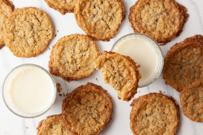 Oatmeal cookies are scattered around two glasses of milk on a white surface. Some cookies are broken, adding to the rustic presentation.