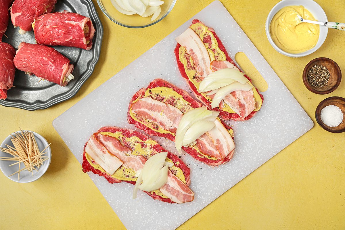 Slices of raw beef topped with mustard, bacon, and onions on a white cutting board. Ingredients, including more beef, sliced onions, mustard, salt, and pepper, surround the board on a yellow surface.