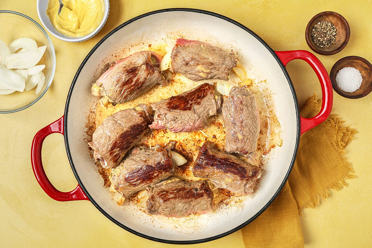 A red-handled skillet on a yellow surface contains browned beef pieces. Nearby are small bowls with sliced onions, mustard, black pepper, and salt. A yellow napkin is partially visible under the skillet.