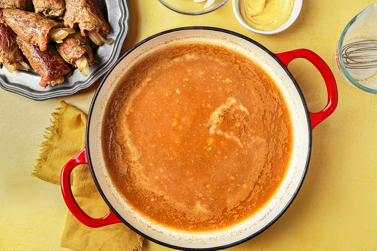 A red pot filled with a creamy brown sauce sits on a yellow surface. Nearby are a plate with cooked meat, a bowl of mustard, a whisk, and a folded yellow napkin. The scene suggests preparation for a meal.