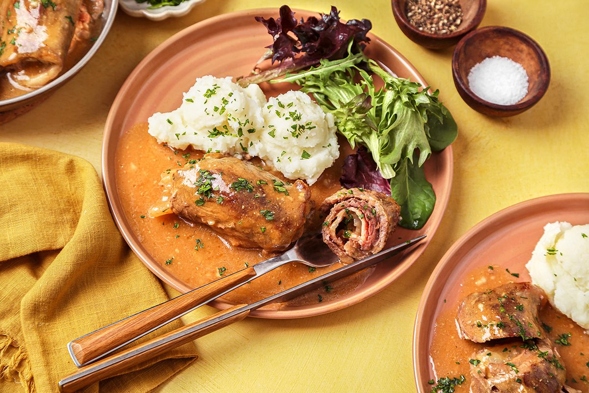 A plate of stuffed cabbage rolls with tomato sauce is served alongside mashed potatoes and mixed greens. A fork and knife are placed on the plate, and small bowls of seasoning and salt are in the background.