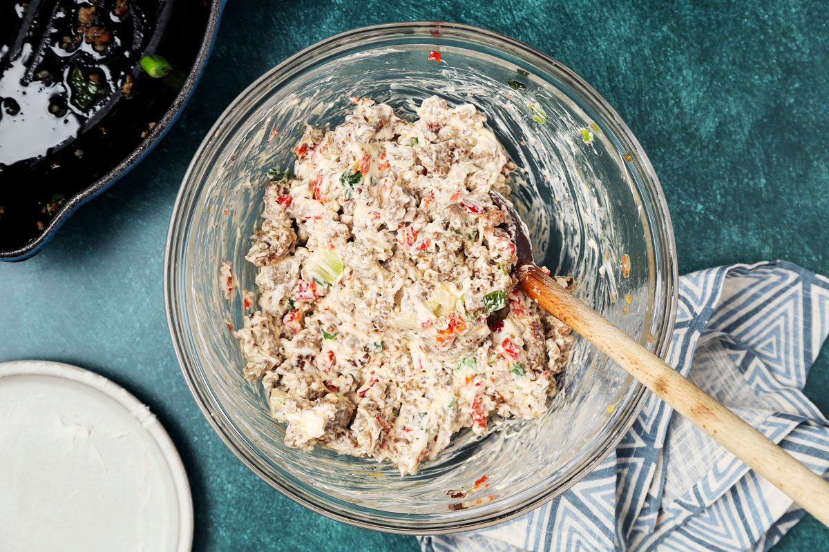 overhead shot of a glass bowl filled with a mixture of ingredients, including sausage, cream cheese, green onions, and red pepper; the bowl is sitting on a teal surface with a wooden spoon resting on the rim, the spoon is also resting on a blue and white patterned dish towel