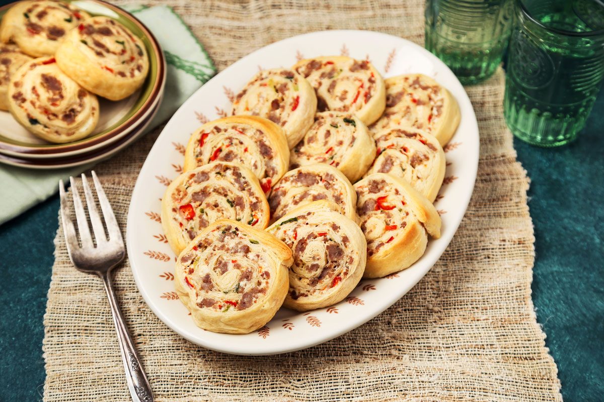 3/4th shot of a plate of sausage rolls; the rolls are arranged in a circle on the plate, which is a white ceramic with a brown and white pattern; the plate is sitting on a brown placemat, which is made of woven straw; a fork is placed next to the plate; in the background, there are two green glass tumblers, one full and the other empty, sitting on a blue surface
