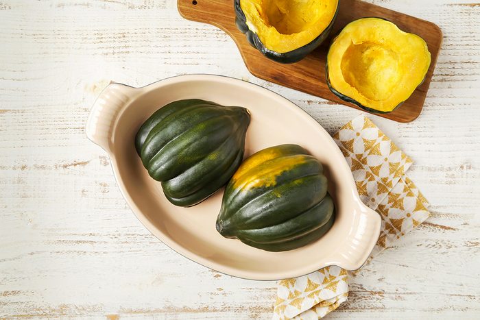 Two whole acorn squashes sit in a cream-colored baking dish on a wooden table. Nearby, two squash halves rest on a wooden board, revealing their orange interiors. A patterned napkin is placed beside the dish.