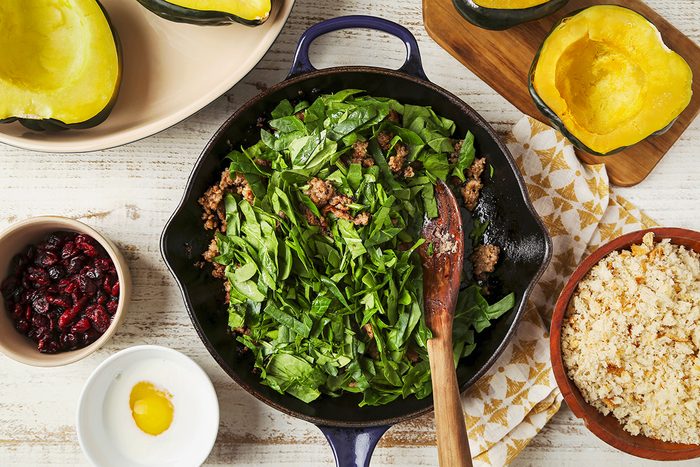 A skillet with spinach and ground meat mixture being stirred by a wooden spoon. Surrounding the skillet are acorn squash halves, a bowl of dried cranberries, a bowl of cooked rice, and a small dish with a cracked egg.