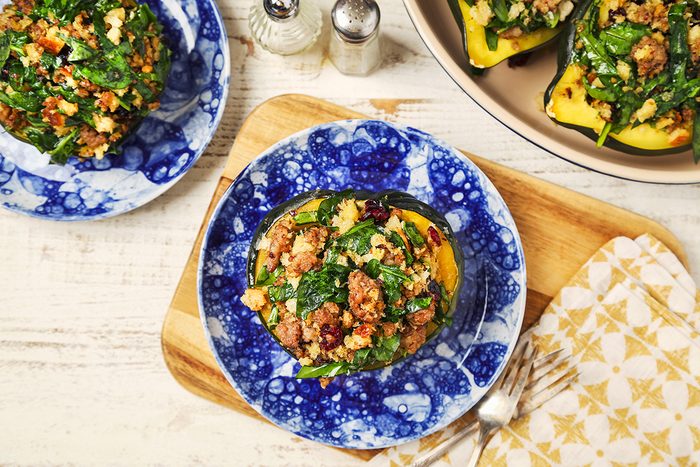 A blue and white plate holds a stuffed acorn squash filled with a mixture of spinach, quinoa, cranberries, and crumbled cheese. It's served on a wooden board with cutlery and a napkin nearby. Other plates with similar dishes are partially visible.