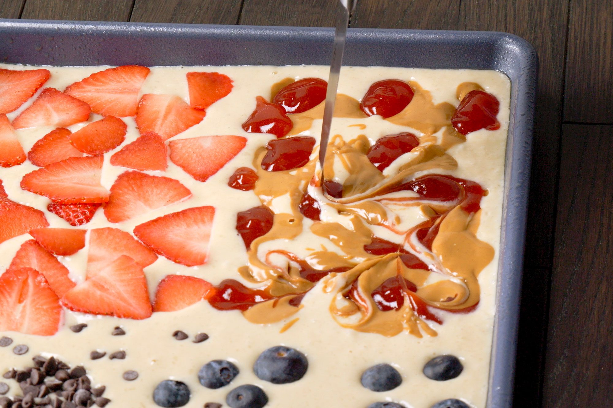 closeup shot of a sheet pan filled with a batter for Sheet Pan Pancakes, arranged with various toppings; sliced strawberries are placed on one side, The middle section features swirls of peanut butter and fruit or berry sauce, On the opposite side, blueberries and chocolate chips are scattered