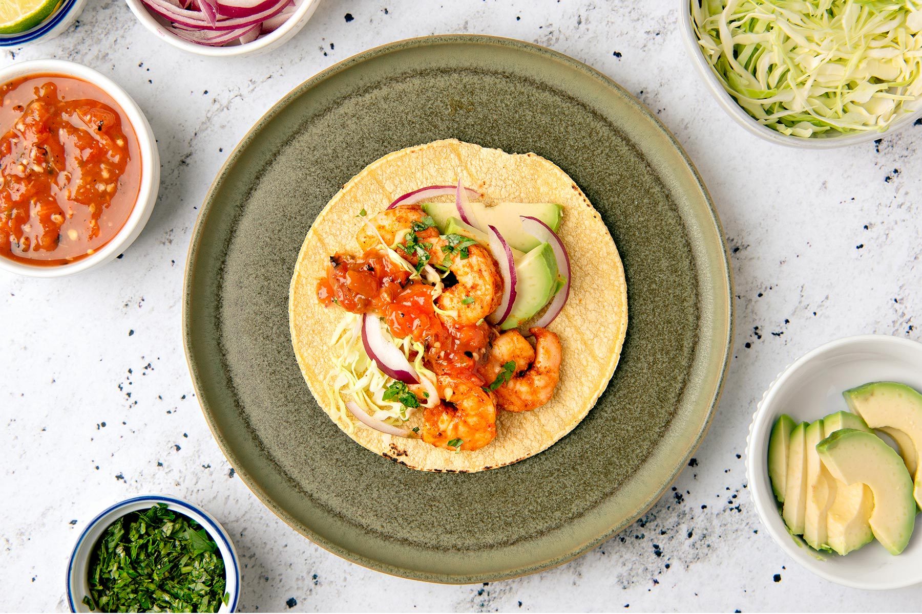 overhead shot of shrimp taco with a variety of toppings, arranged on a green plate, surrounded by various ingredients; the taco is filled with seasoned shrimp, chopped cabbage, red onion, chopped avocado, and a salsa-like sauce; there is also a small bowl of chopped cilantro to the left of the taco, and a bowl of sliced avocado to the right; in the top left corner of the image, there are small bowls of lime wedges and a red salsa; in the top right corner of the image, there is a bowl of shredded cabbage