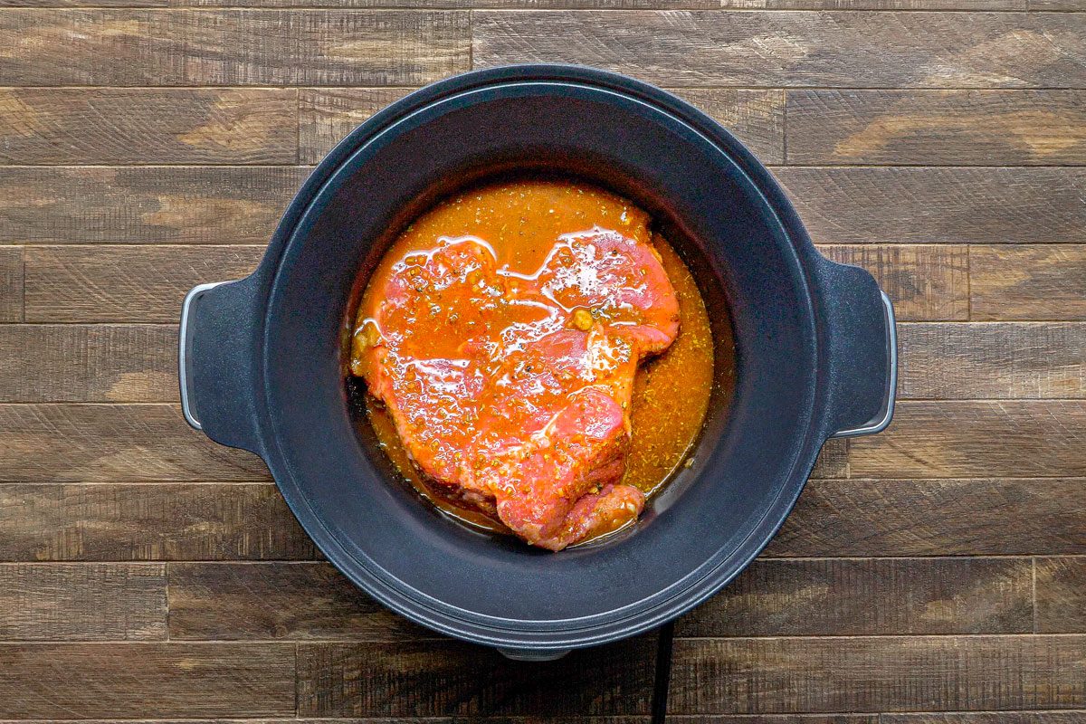 overhead shot of a raw piece of meat in a black slow cooker on a wooden surface; the meat is covered in a red marinade and sits in a pool of the marinade at the bottom of the slow cooker