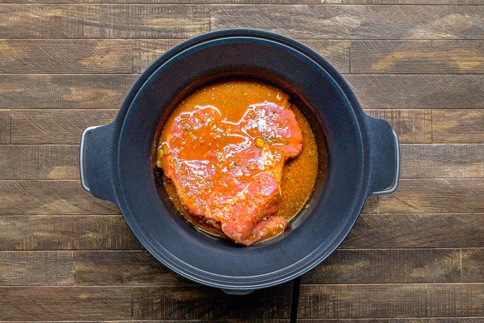 overhead shot of a raw piece of meat in a black slow cooker on a wooden surface; the meat is covered in a red marinade and sits in a pool of the marinade at the bottom of the slow cooker