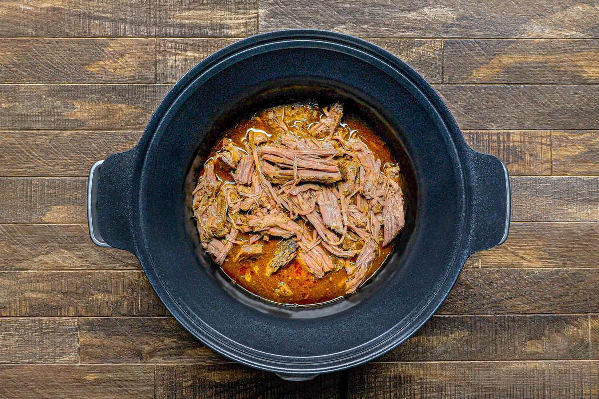 overhead shot of a black slow cooker filled with shredded meat in a brown sauce, it is set on a wooden surface