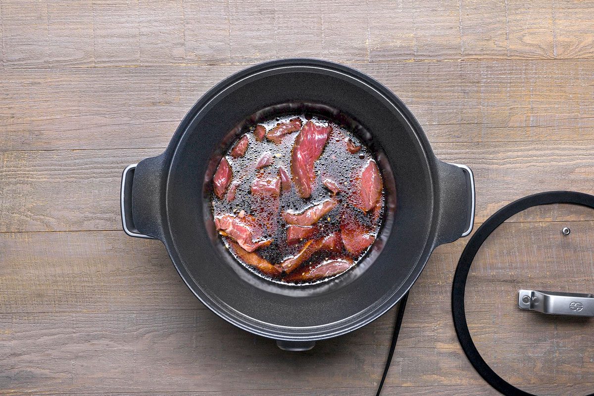 overhead shot of a black slow cooker with a glass lid on a wooden surface; the slow cooker is filled with raw meat submerged in a dark liquid