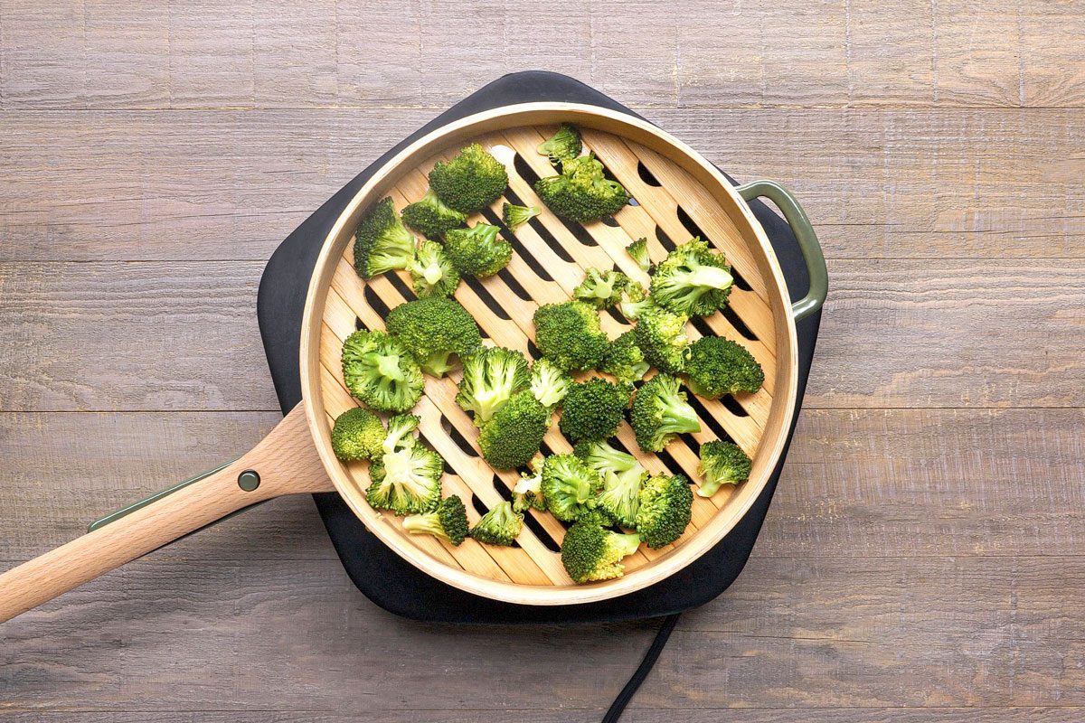 overhead shot of a bamboo steamer filled with broccoli florets; the steamer is sitting on a black base, and the whole setup is on a wooden table