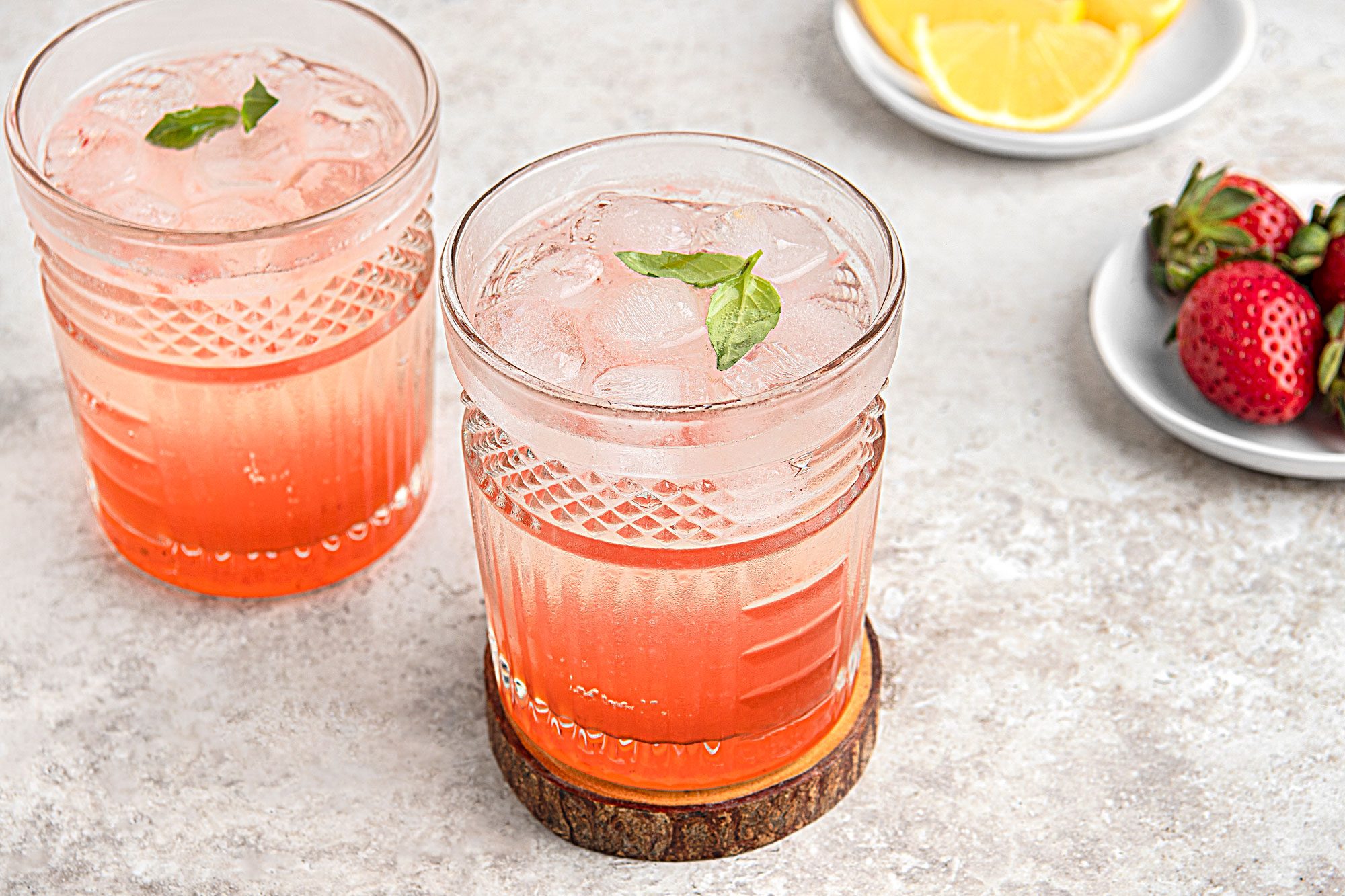 Close view shot of Strawberry-Basil Mocktail; served into rock glasses filled with ice; Top with club soda and additional basil leaves; a glass on wood coaster; strawberry on plates; lime slices on another plate; grey surface;
