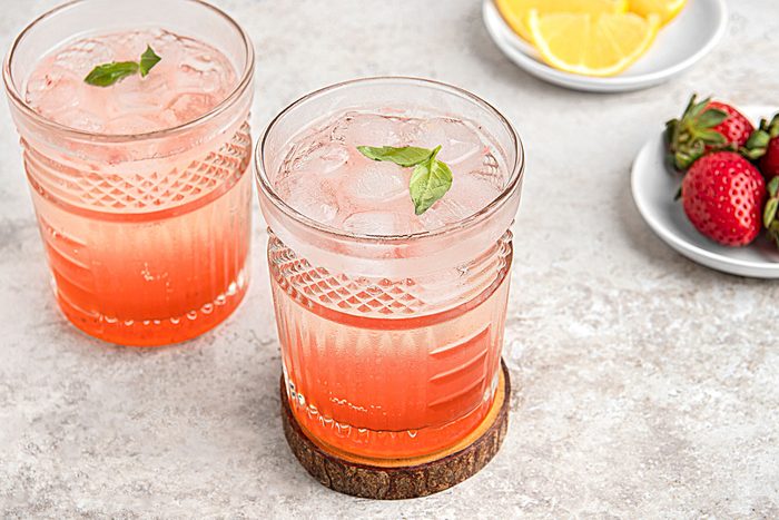 Close view shot of Strawberry-Basil Mocktail; served into rock glasses filled with ice; Top with club soda and additional basil leaves; a glass on wood coaster; strawberry on plates; lime slices on another plate; grey surface;