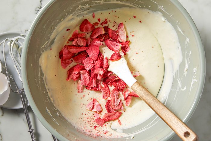 Overhead view of mixing the cream cheese mixture with freeze-dried strawberries using a wooden spatula in a large bowl. Next to it is a used whisk.