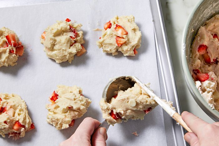 Overhead view of scooping the cookie dough onto a parchment paper-lined baking sheet using the help of a spatula. Next to it lies the large bowl containing the cookie dough.