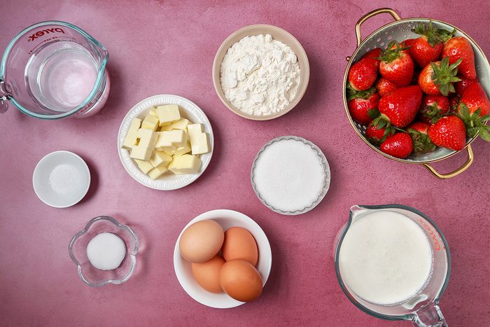 Overhead view of ingredients in containers required to make strawberry cream puffs kept on a pink tabletop.