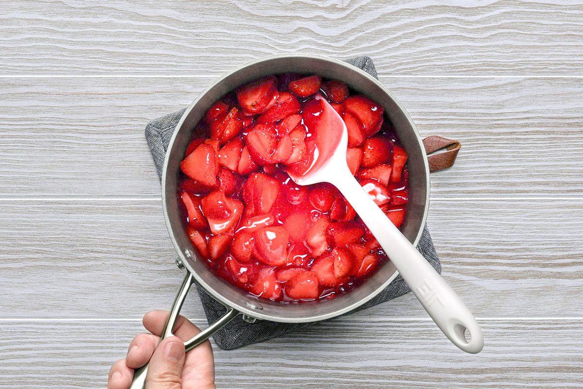 overhead shot of a pot of cooked strawberries on a white wooden table; the strawberries are cooked in their own juices and are a bright red color; a white spatula is in the pot;