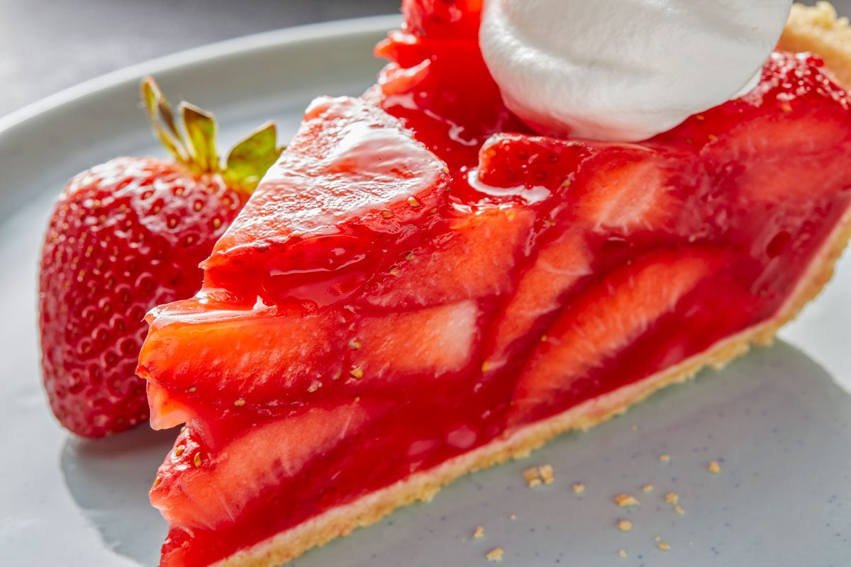 closeup shot of slice of a strawberry pie topped with glossy red strawberry glaze; beside the slice, a whole strawberry is there, and a dollop of whipped cream sits on top