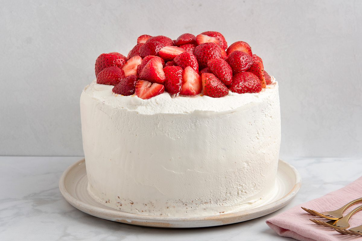 A white frosted cake topped with fresh, halved strawberries on a white plate. A pink napkin and two forks are on the right side. The background is a light gray marble surface.