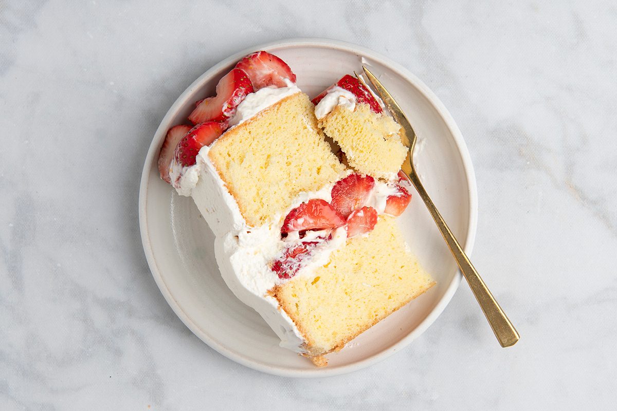 A slice of vanilla cake with layers of whipped cream and fresh strawberries on a white plate, accompanied by a gold fork, set on a marble surface.