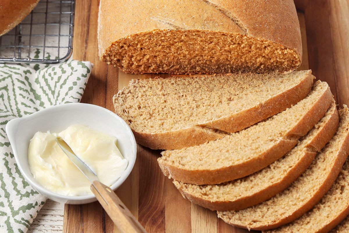 closeup shot of Swedish Limpa Bread on a wooden cutting board, to the left of the bread, there's a small white bowl containing butter and a butter knife with a wooden handle, the background consists of white wooden planks