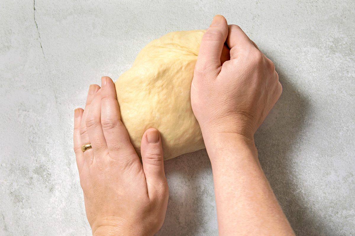 Hands kneading a piece of dough on a light, speckled countertop. One hand holds the dough steady while the other presses into it, showing the kneading process. 