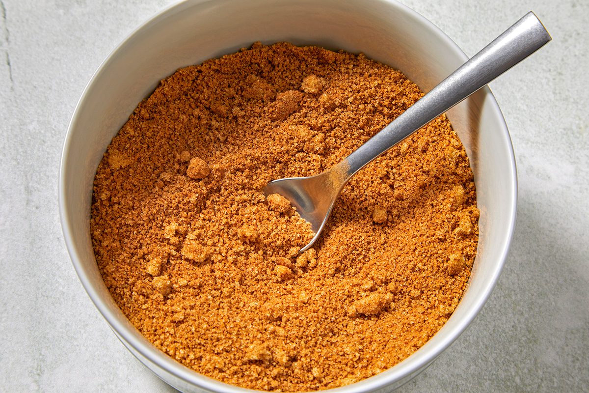 A white bowl filled with golden-brown graham cracker crumbs. A metal spoon rests inside the bowl, partially buried in the crumbs. The bowl is placed on a light gray surface.