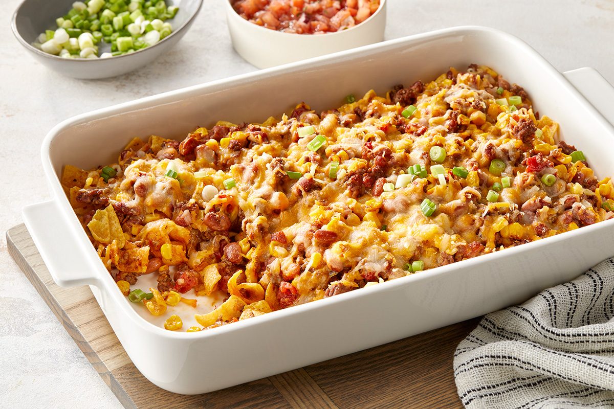 A baked casserole in a white dish on a wooden board. It's topped with melted cheese, ground meat, and sliced green onions. In the background are small bowls of chopped green onions and diced tomatoes. A striped cloth is partially visible.