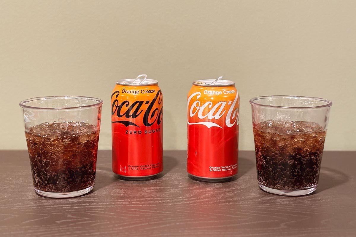 Can of Coca Cola Orange Cream Zero sugar next to an iced glass of cola on the left; Can of Coca Cola Orange Cream with cola in an iced glass on the right, on a dark wooden table and beige background