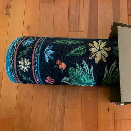 Black rug with floral and leaf design rolled up emerging from a box on a wood floor