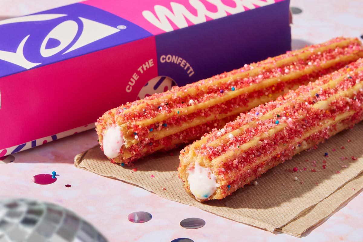 Two Taco Bell x Milk Bar stuffed churros coated with pink sprinkles sit on a brown napkin in front of a pink and purple box
