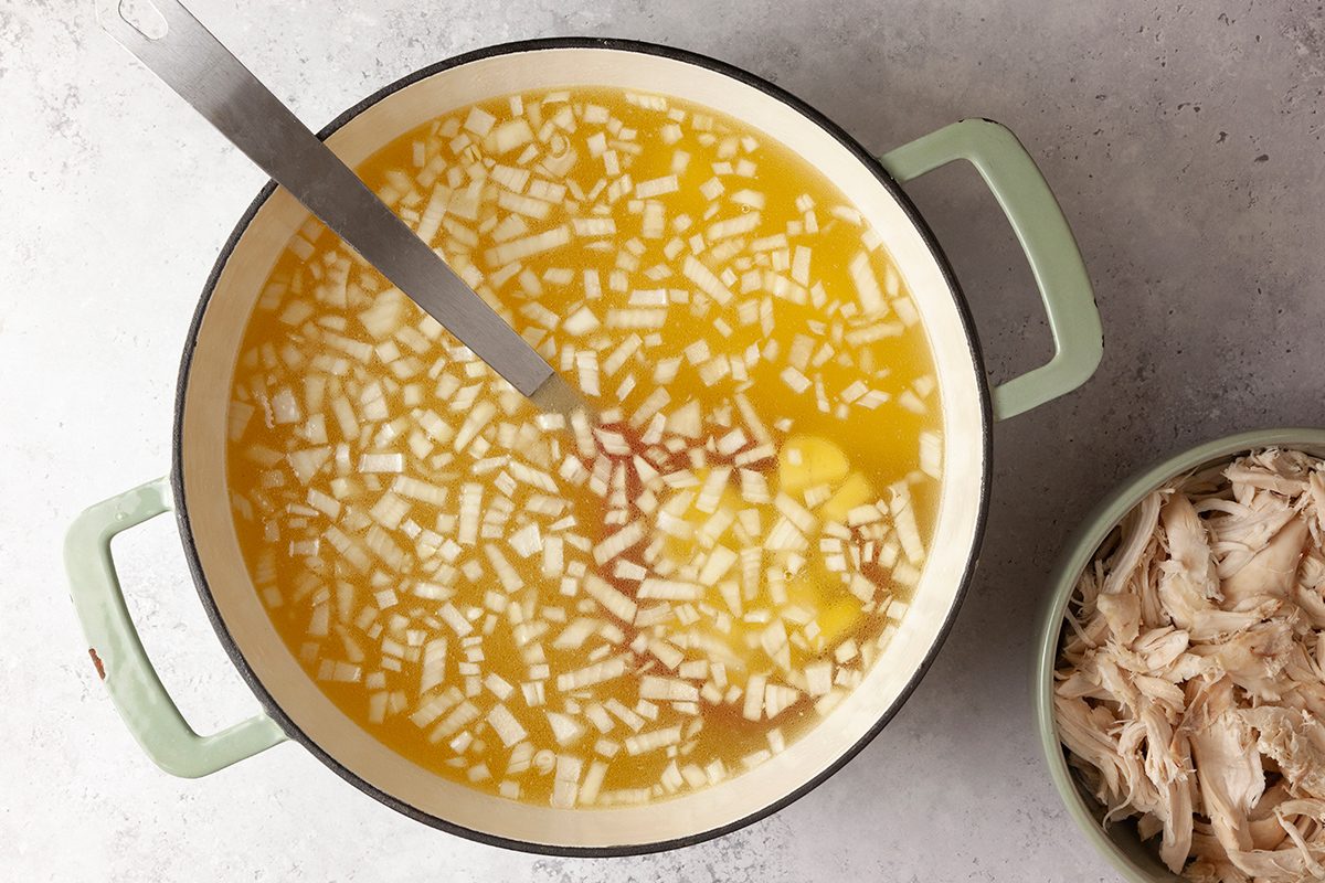A pot of yellow broth with chopped vegetables and a ladle inside, placed on a gray surface. Next to it is a bowl filled with shredded chicken.