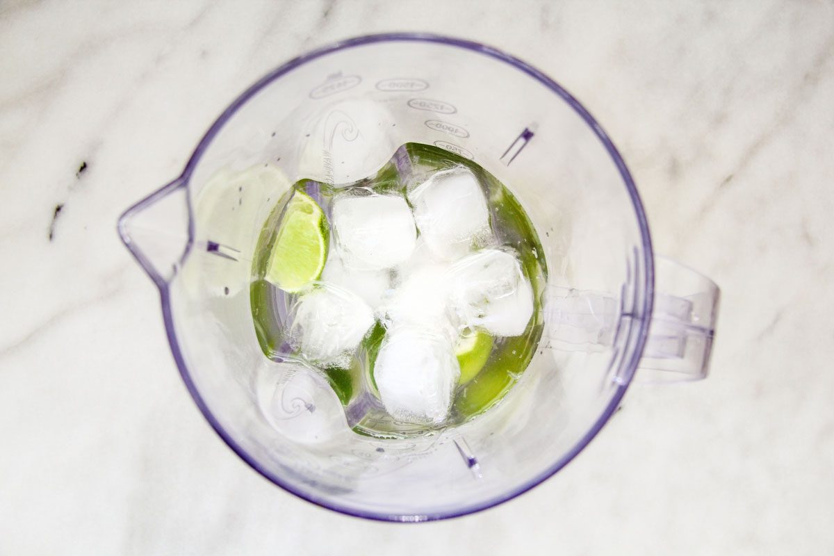 A blender pitcher filled with lime wedges, ice cubes, and a liquid, viewed from above on a light marble surface.