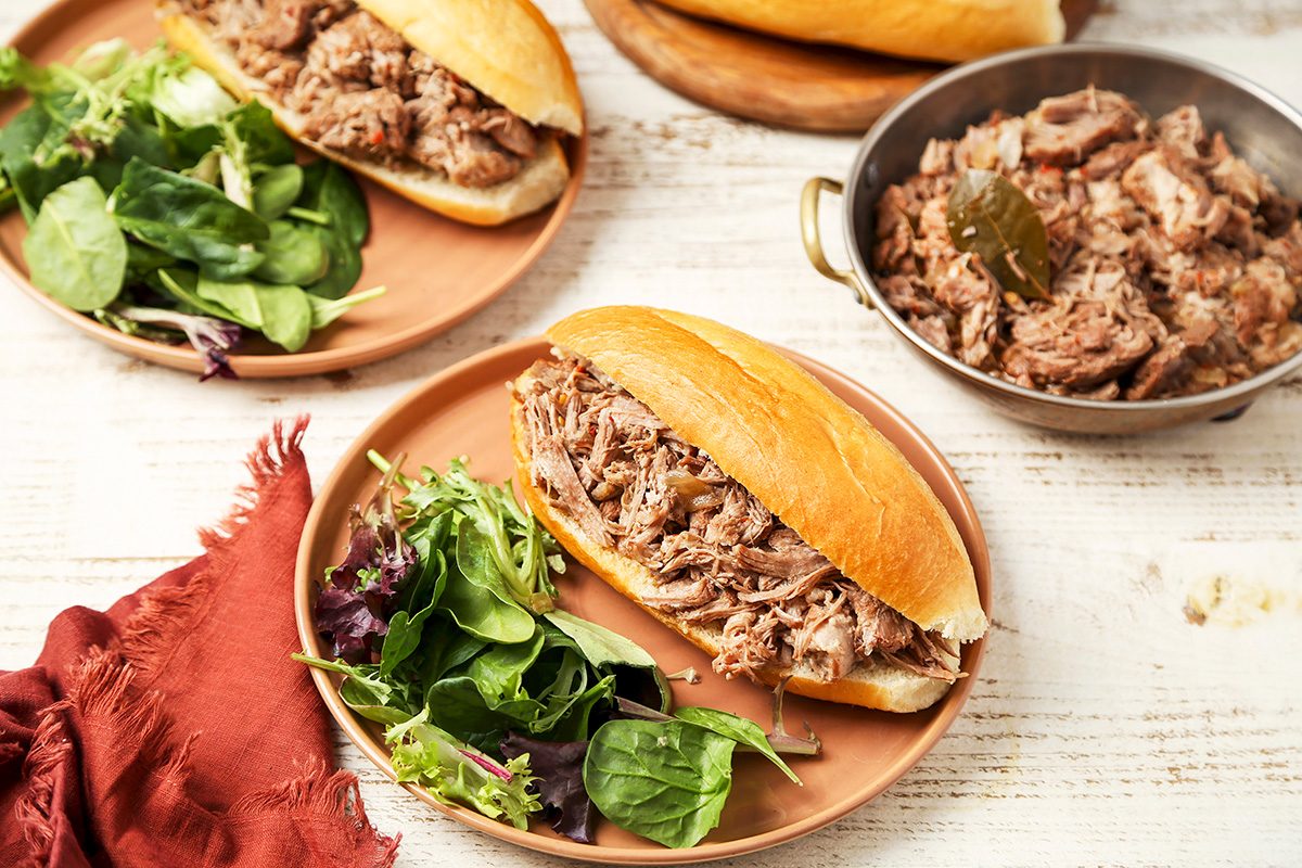 Two pulled pork sandwiches on hoagie rolls are served with mixed greens on a wooden table. A pot of shredded pork is nearby, along with a red cloth napkin.